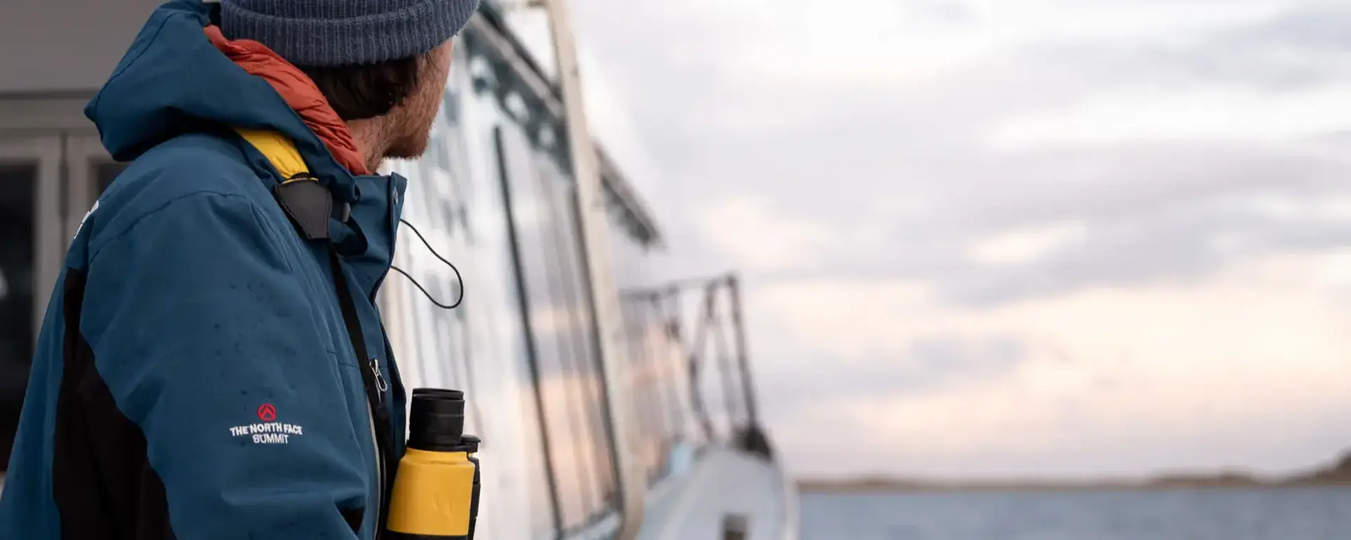 A man standing outside on a modern catamaran while it cruises across calm waters looks out ahead. 
