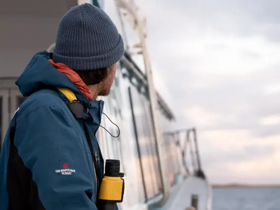 A man standing outside on a modern catamaran while it cruises across calm waters looks out ahead. 