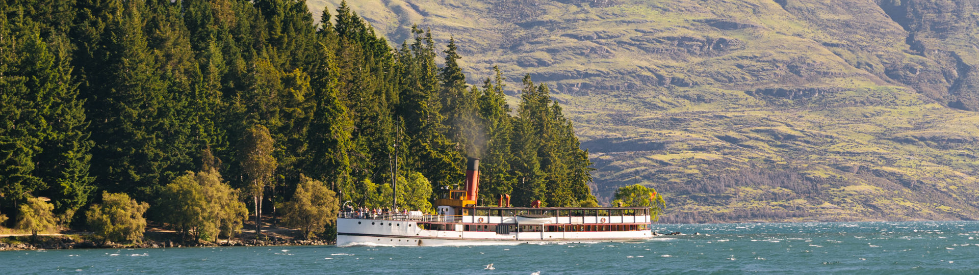 The historic TSS Earnslaw vintage steamship cruises past Queenstown Gardens with Cecil Peak mountain in the background.