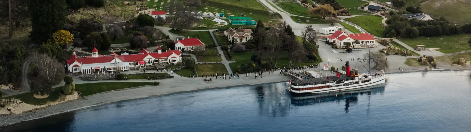 Aerial drone shot of the lakeside Walter Peak High Country Farm with the historic TSS Earnslaw steamship docked at the wharf. 