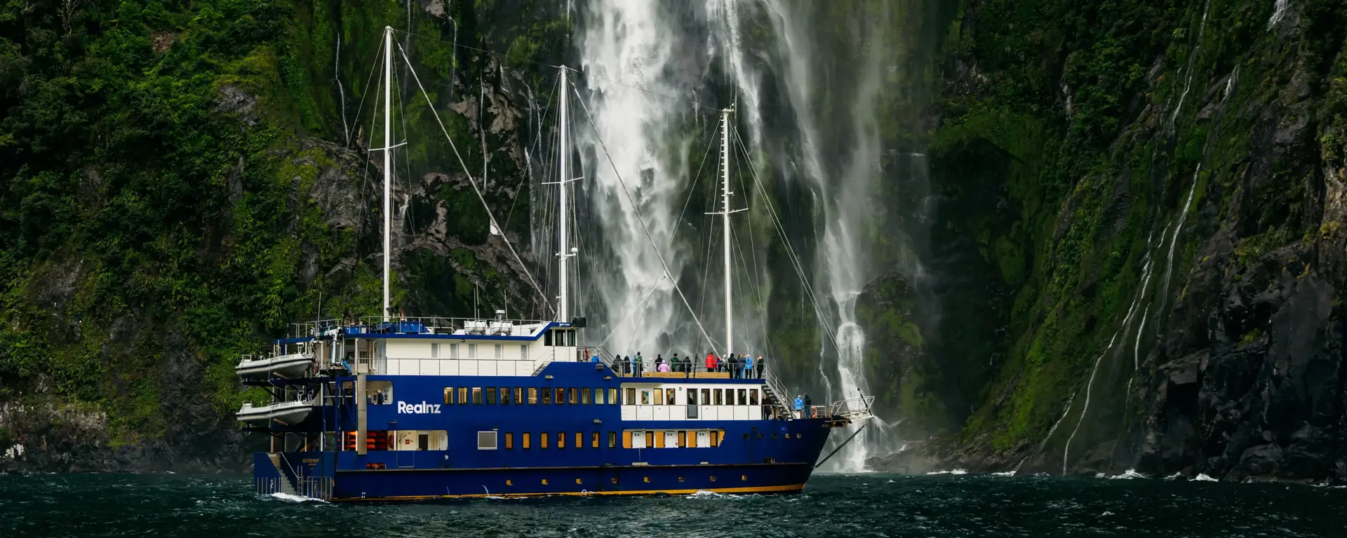 Cruise vessel beneath a towering waterfall in Milford Sound.