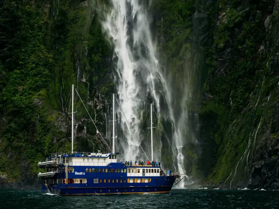 Cruise vessel beneath a towering waterfall in Milford Sound.