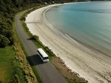 RealNZ coach travelling along a coastal road beside a sandy beach.