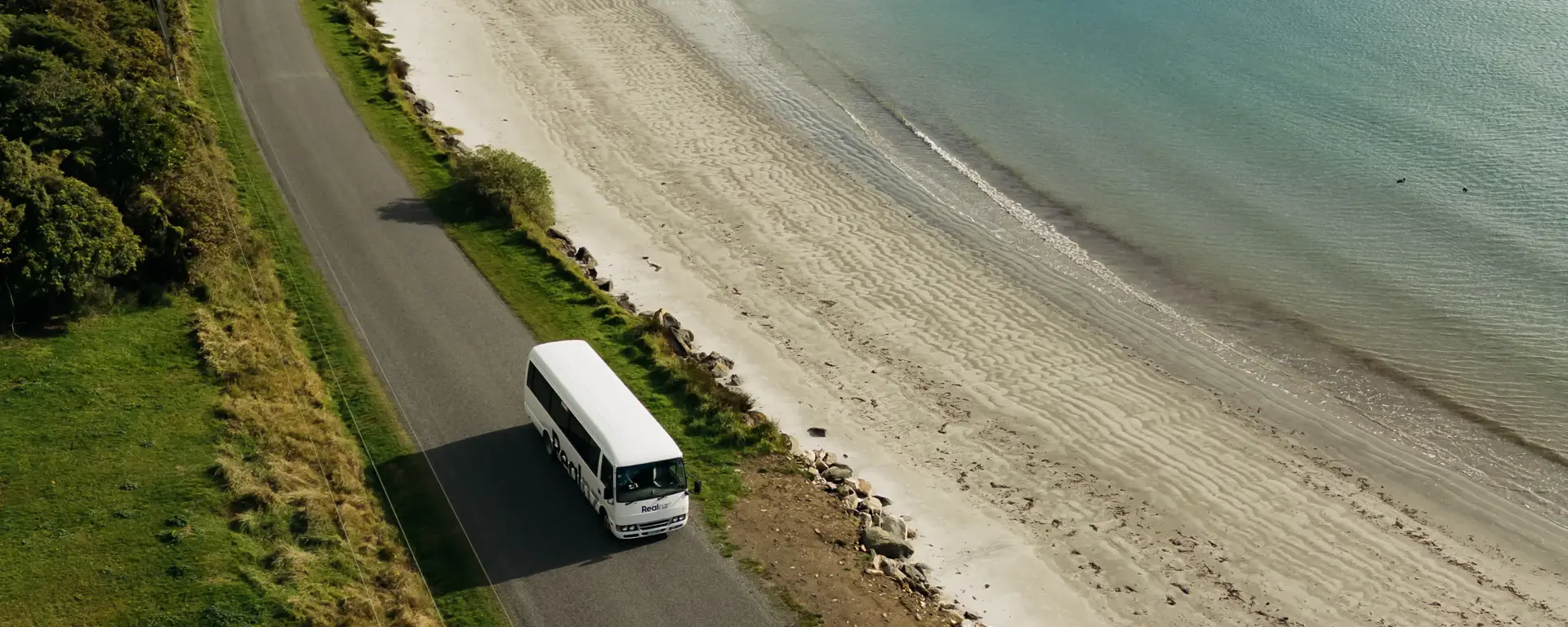 RealNZ coach travelling along a coastal road beside a sandy beach.
