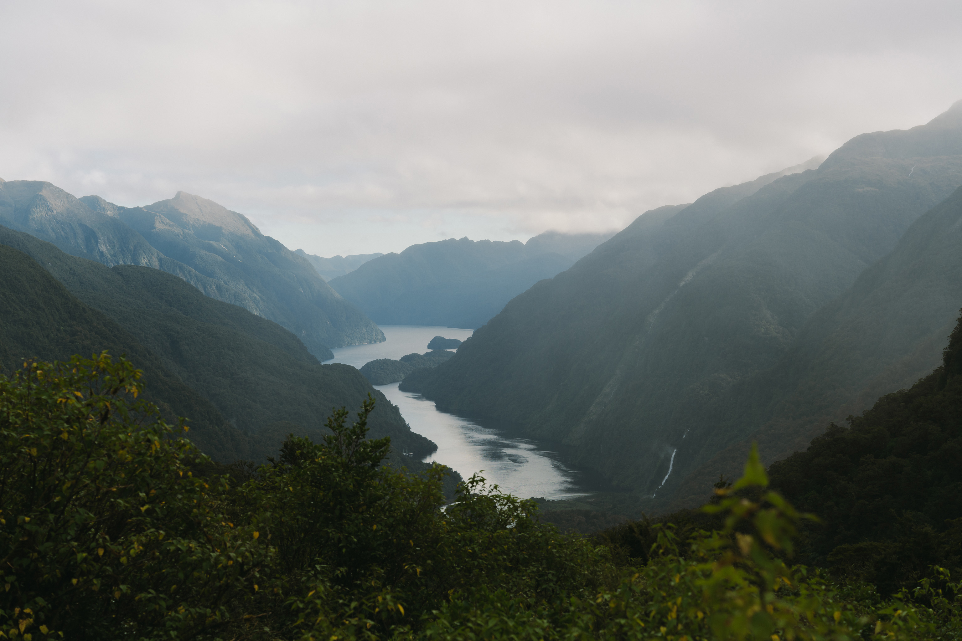 A misty fjord valley with steep forested mountains on either side, a calm winding waterway stretching into the distance, and a small waterfall visible on the right slope under an overcast sky.