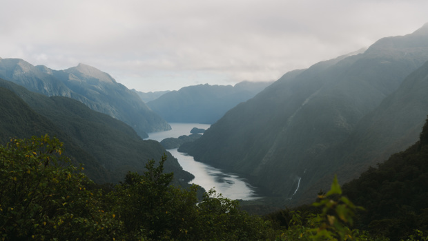 A misty fjord valley with steep forested mountains on either side, a calm winding waterway stretching into the distance, and a small waterfall visible on the right slope under an overcast sky.