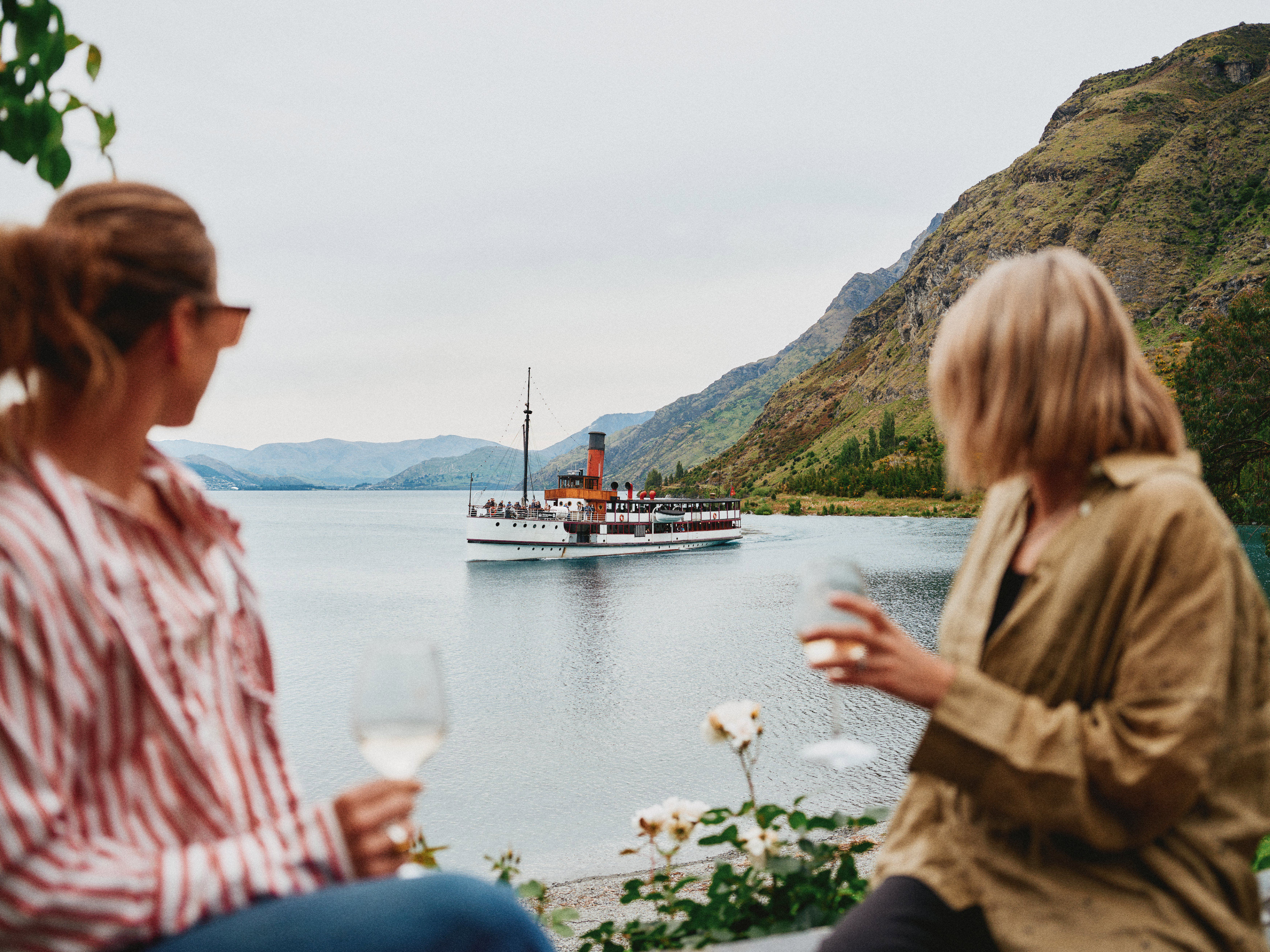Two women enjoying a glass of wine sitting lakeside outdoors look out towards the TSS Earnslaw cruising on Lake Whatkatipu.