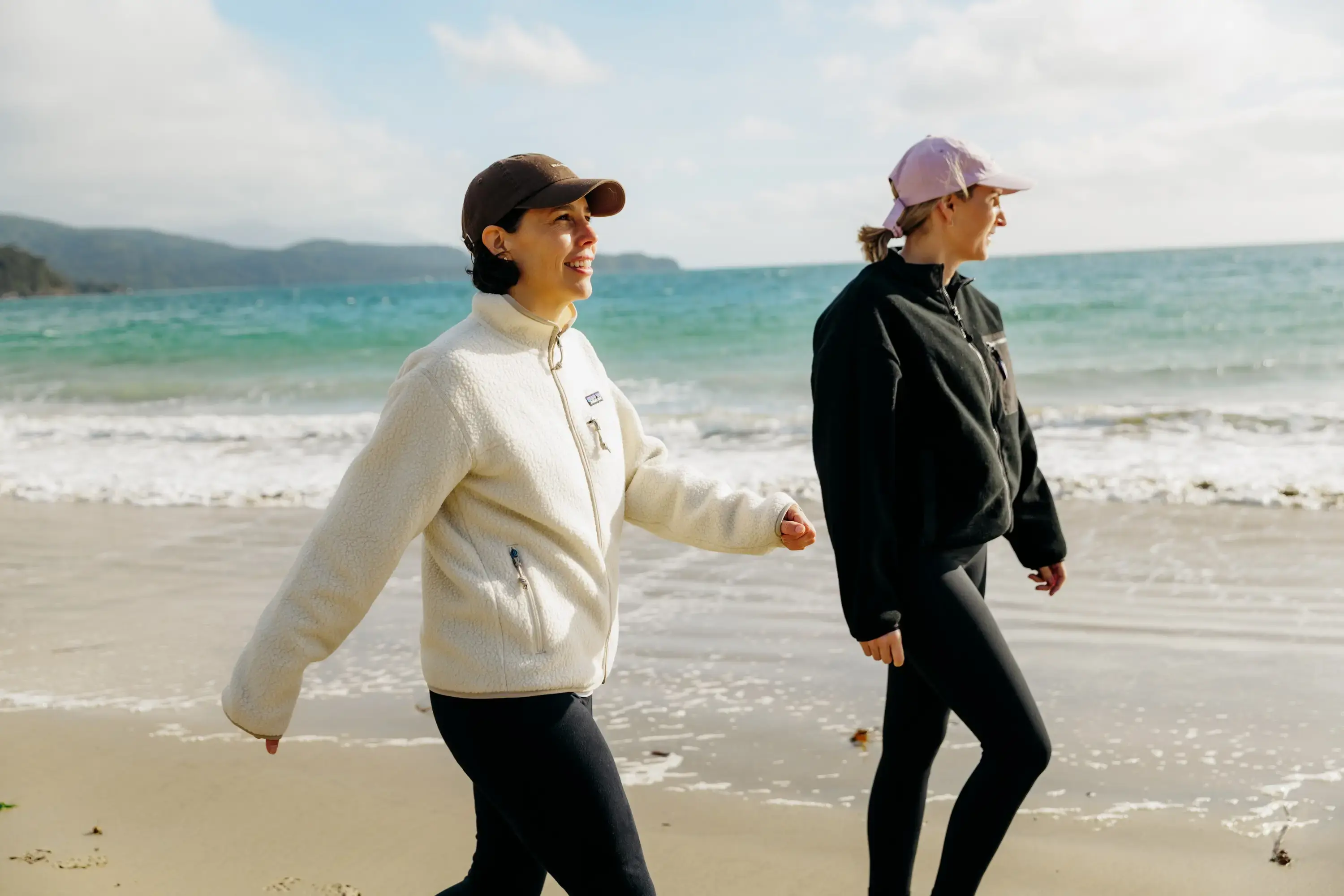 Two women dressed in leggings and fleeces walk along a golden beach with blue ocean in the background. 