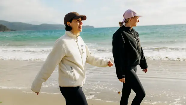 Two women dressed in leggings and fleeces walk along a golden beach with blue ocean in the background. 