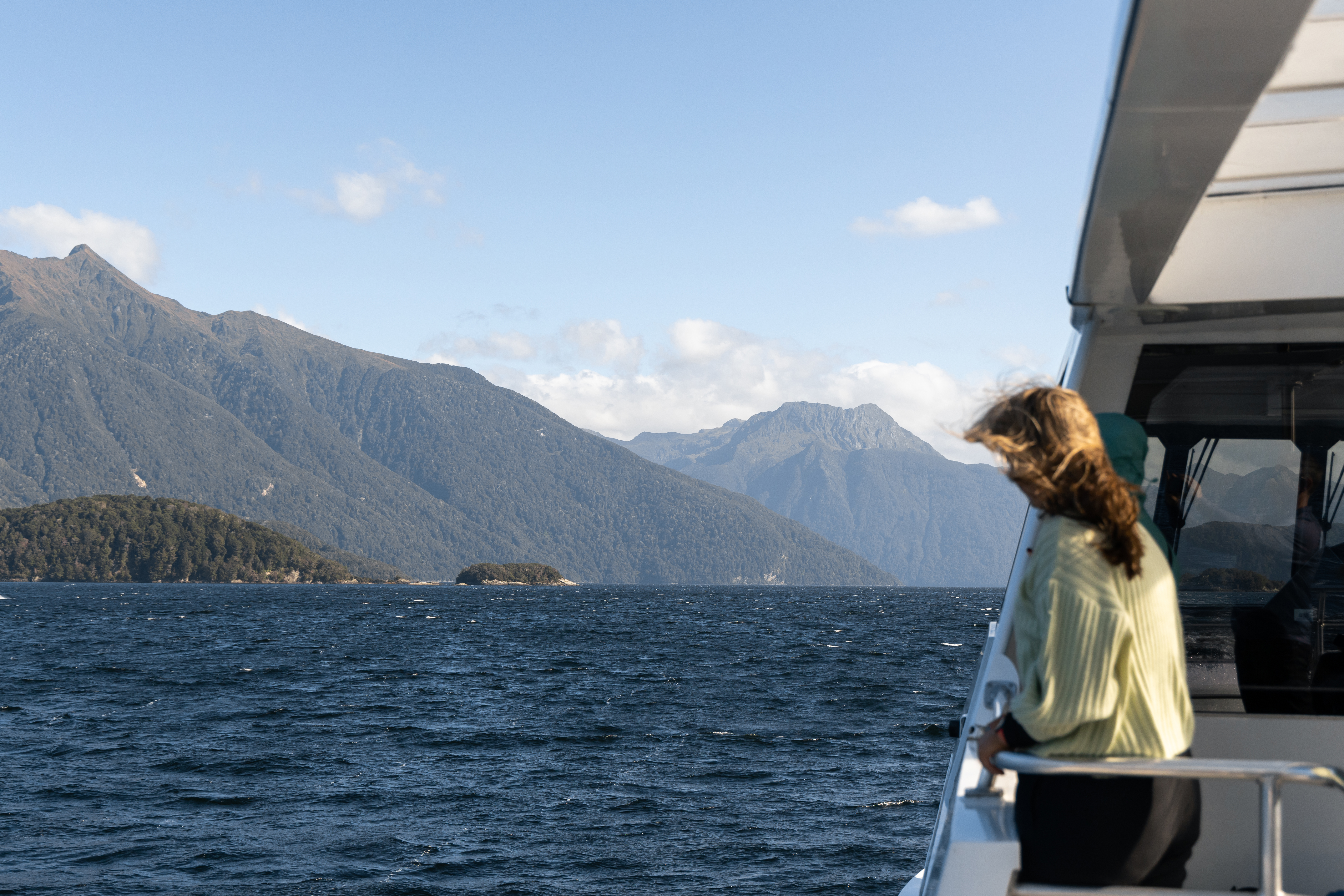 A women stands on an outside deck of a cruise boat looking out at the view of a blue lake surrounded by tree covered mountains.