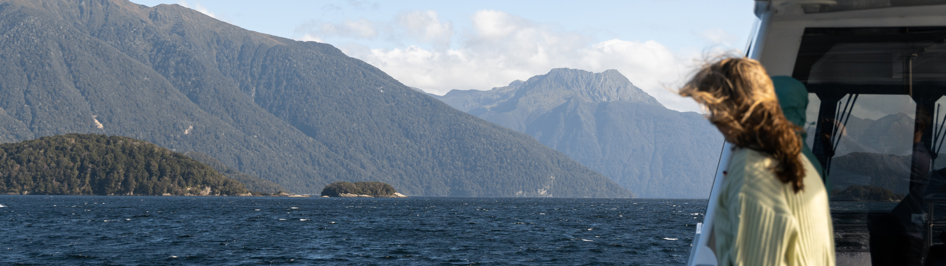 A women stands on an outside deck of a cruise boat looking out at the view of a blue lake surrounded by tree covered mountains.
