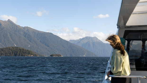 A women stands on an outside deck of a cruise boat looking out at the view of a blue lake surrounded by tree covered mountains.