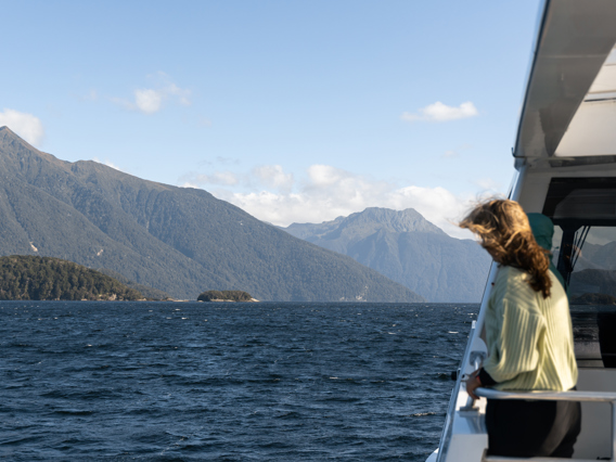 A women stands on an outside deck of a cruise boat looking out at the view of a blue lake surrounded by tree covered mountains.