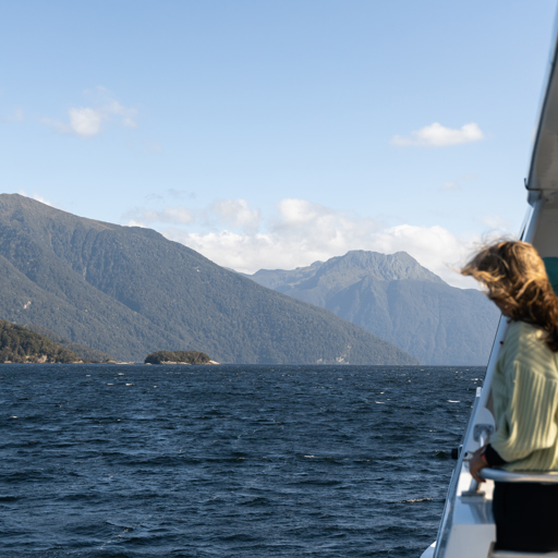 A women stands on an outside deck of a cruise boat looking out at the view of a blue lake surrounded by tree covered mountains.