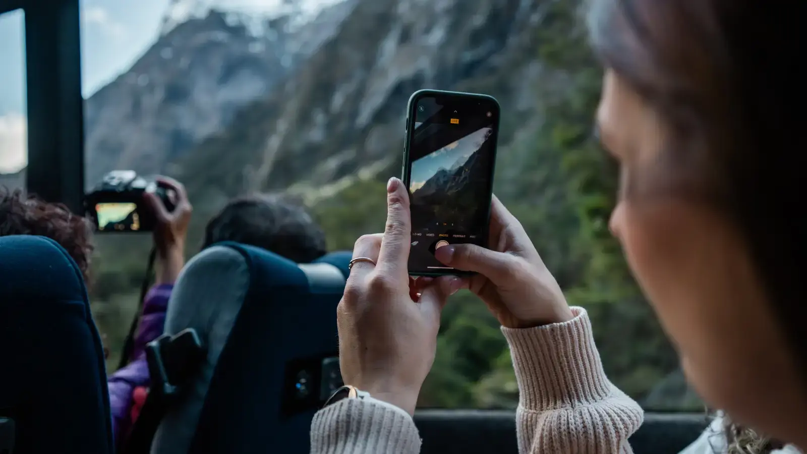 A passenger on a bus photographs the mountainous scenery through the window while another traveler uses a camera nearby