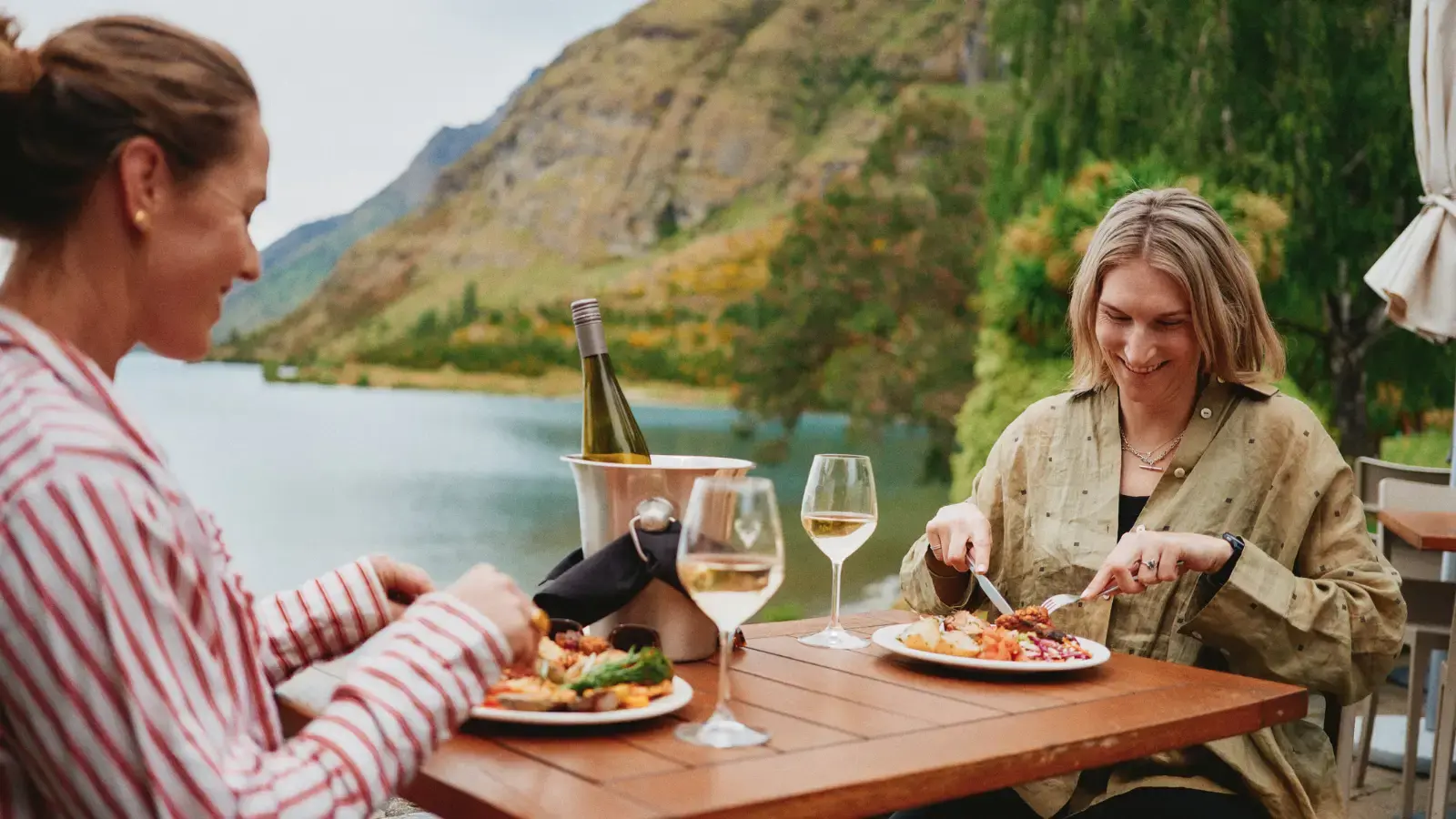 Two women enjoy an outdoor meal of BBQed meats, salads, vegetables and a bottle of wine, while sittnig lakeside. 