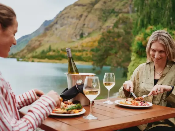 Two women enjoy an outdoor meal of BBQed meats, salads, vegetables and a bottle of wine, while sittnig lakeside. 