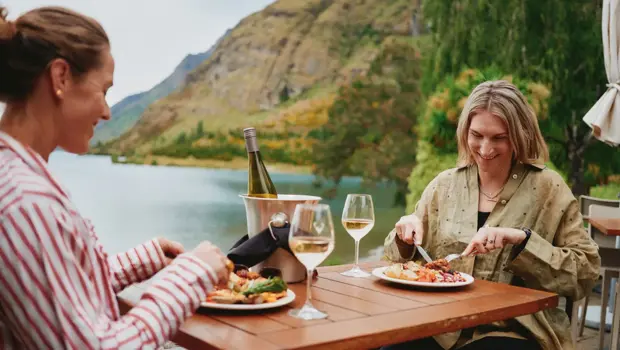 Two women enjoy an outdoor meal of BBQed meats, salads, vegetables and a bottle of wine, while sittnig lakeside. 