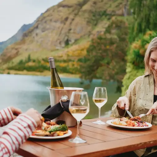 Two women enjoy an outdoor meal of BBQed meats, salads, vegetables and a bottle of wine, while sittnig lakeside. 