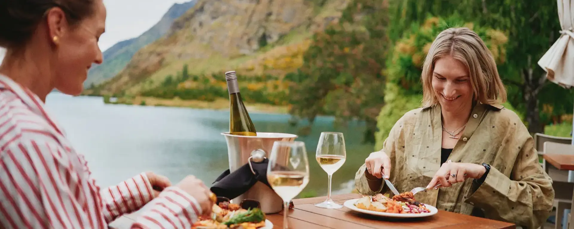 Two women enjoy an outdoor meal of BBQed meats, salads, vegetables and a bottle of wine, while sittnig lakeside. 