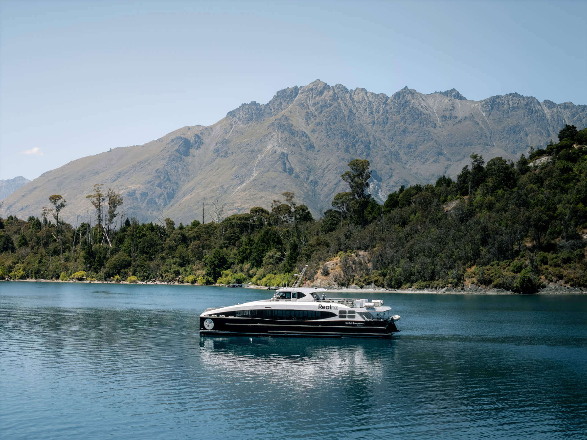 Spirit of Queenstown cruise boat gliding on Lake Wakatipu with rugged mountain peaks and lush greenery in the background on a sunny day.