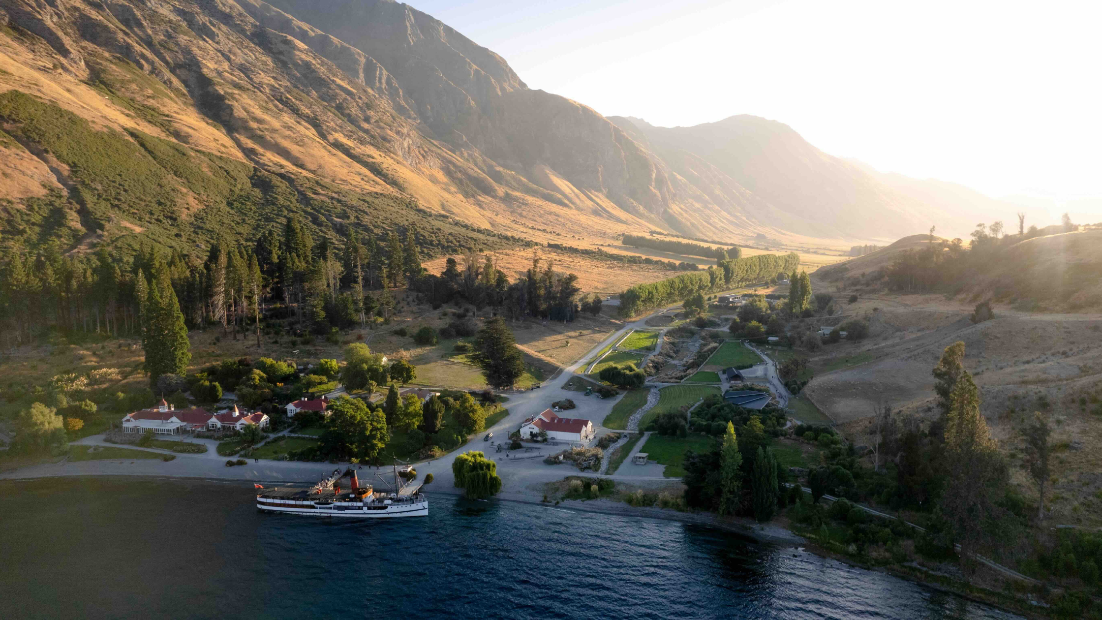 Aerial view of Walter Peak farm buildings on Lake Wakatipu, with mountains in warm evening light.