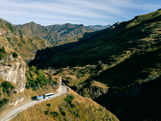 A coach travelling a winding mountain road through the tawny alpine landscape above Queenstown, with rugged peaks stretching into the distance.