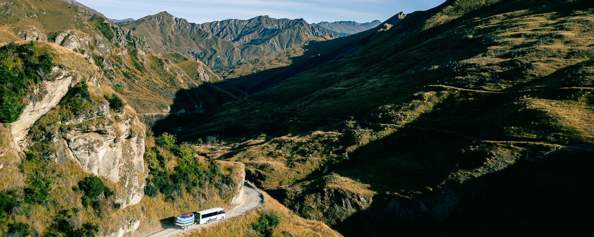 A coach travelling a winding mountain road through the tawny alpine landscape above Queenstown, with rugged peaks stretching into the distance.