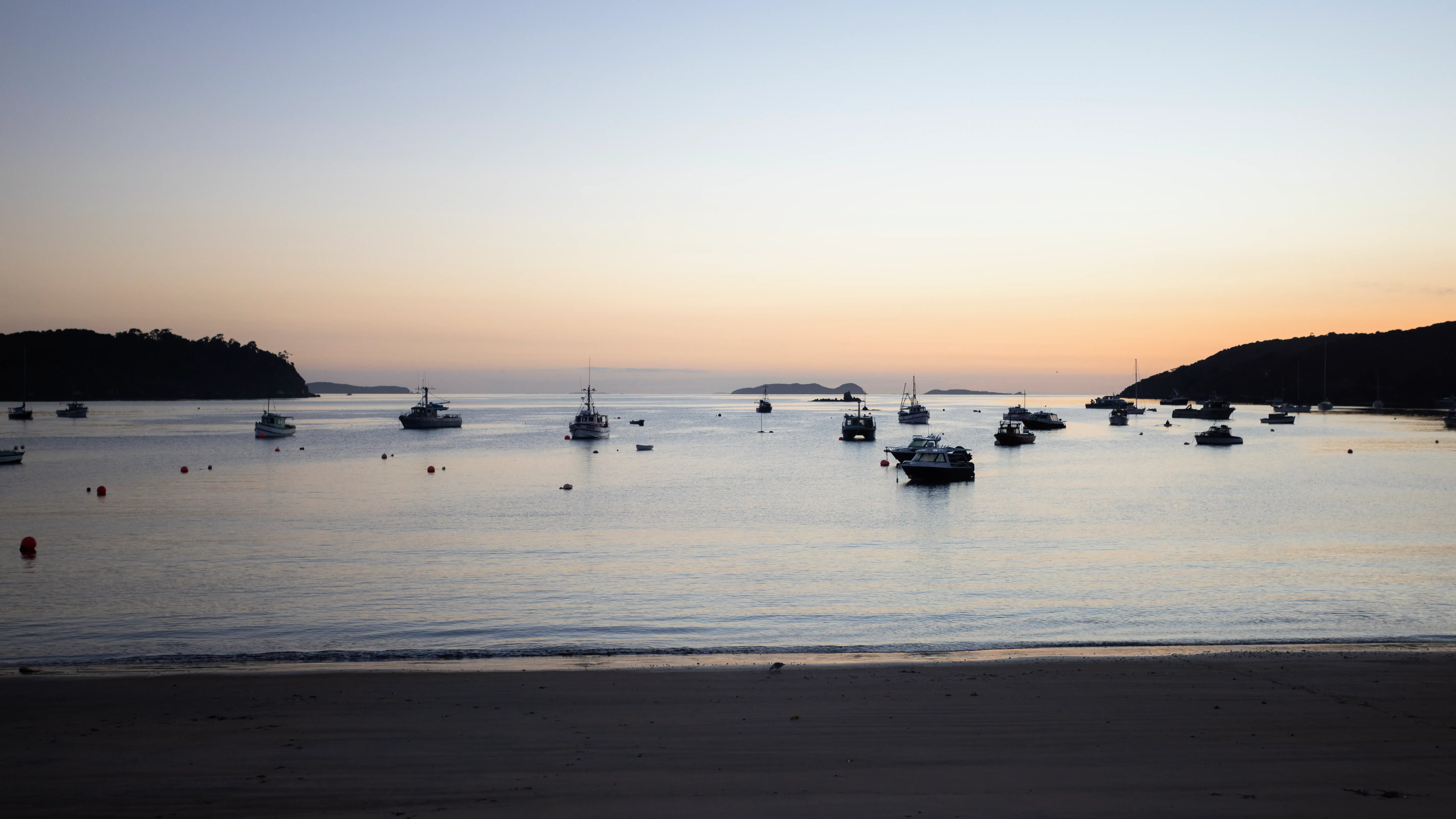 Calm coastal bay at golden hour with sailboats in the distance.
