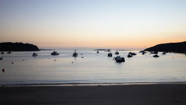 Calm coastal bay at golden hour with sailboats in the distance.