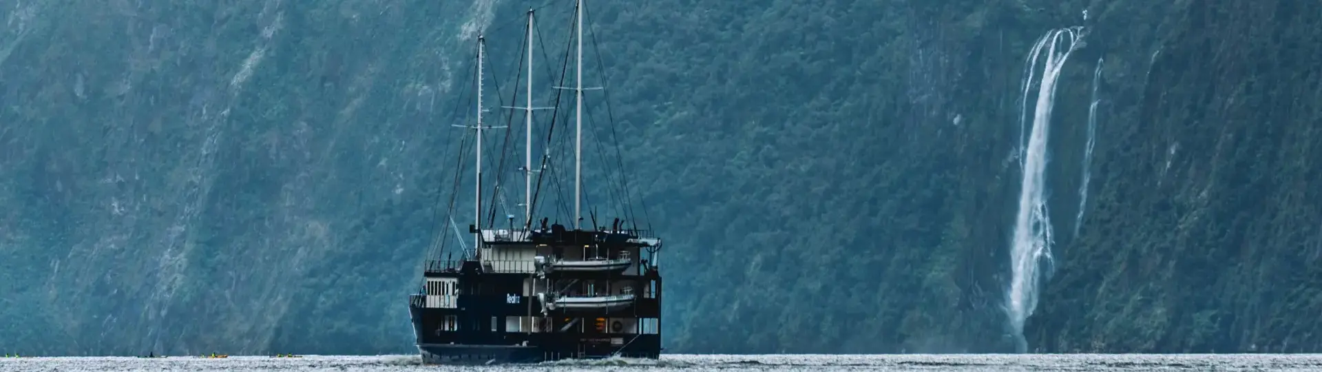 A RealNZ branded tourism overnight boat sails past a waterfall in calm waters in Milford Sound.