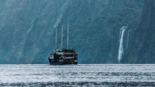 A RealNZ branded tourism overnight boat sails past a waterfall in calm waters in Milford Sound.
