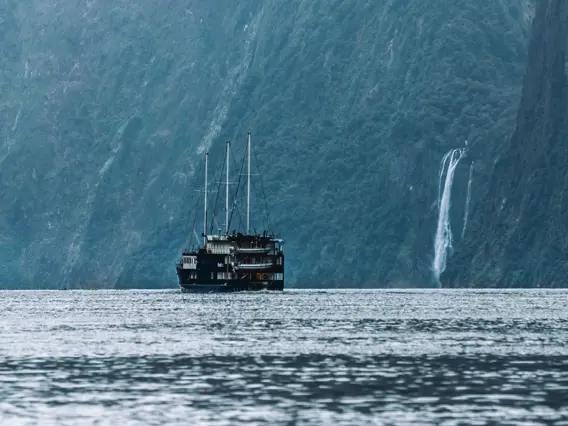 A RealNZ branded tourism overnight boat sails past a waterfall in calm waters in Milford Sound.