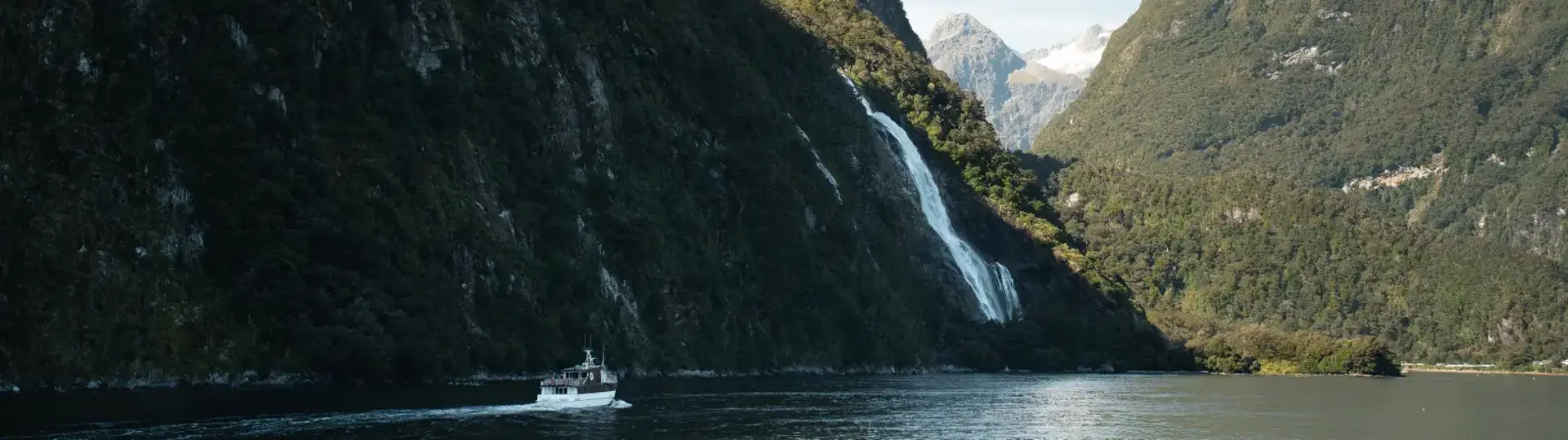 A RealNZ boat cruises through Milford Sound Fiordland towards a gushing waterfall on a blue sky day.