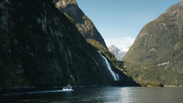 A RealNZ boat cruises through Milford Sound Fiordland towards a gushing waterfall on a blue sky day.