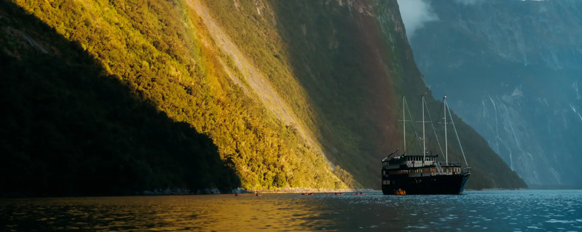 Cruise vessel sailing through dramatic sunlit mountains in Milford Sound.