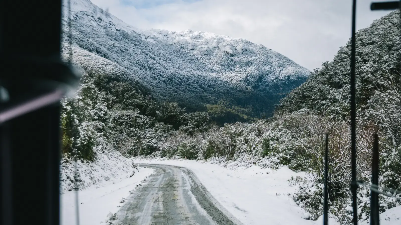 View from inside a coach bus of a wintery, snow-covered track road surrounded by snow-capped mountains. 