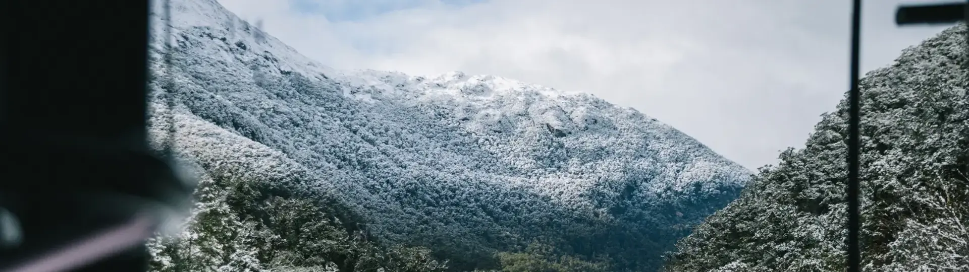 View from inside a coach bus of a wintery, snow-covered track road surrounded by snow-capped mountains. 