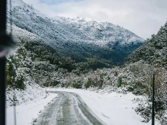 View from inside a coach bus of a wintery, snow-covered track road surrounded by snow-capped mountains. 