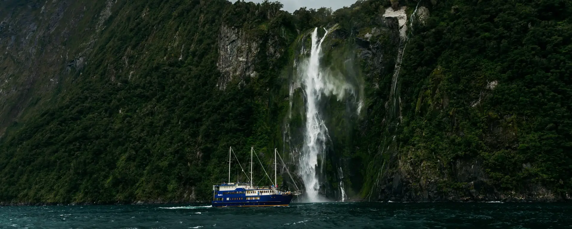 Boat cruising beneath a towering waterfall in Milford Sound.