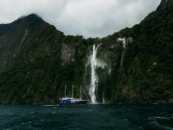 Boat cruising beneath a towering waterfall in Milford Sound.