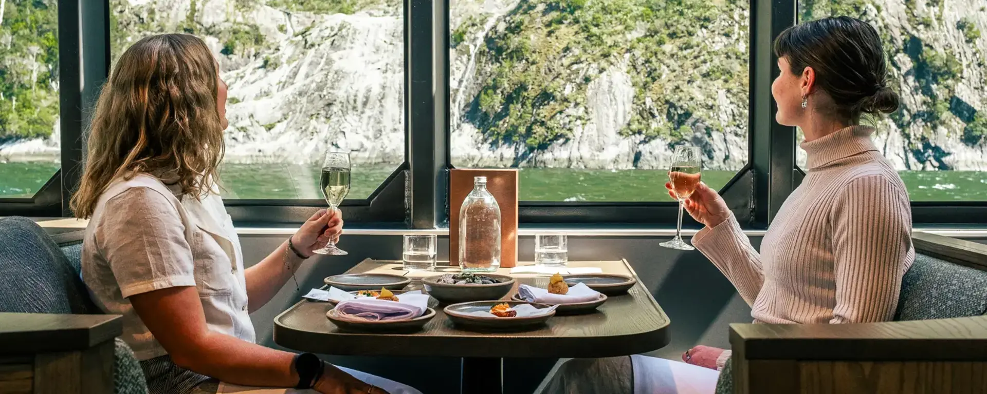 Two guests dining beside large windows overlooking Milford Sound.
