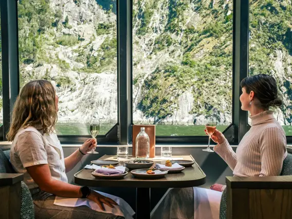 Two guests dining beside large windows overlooking Milford Sound.