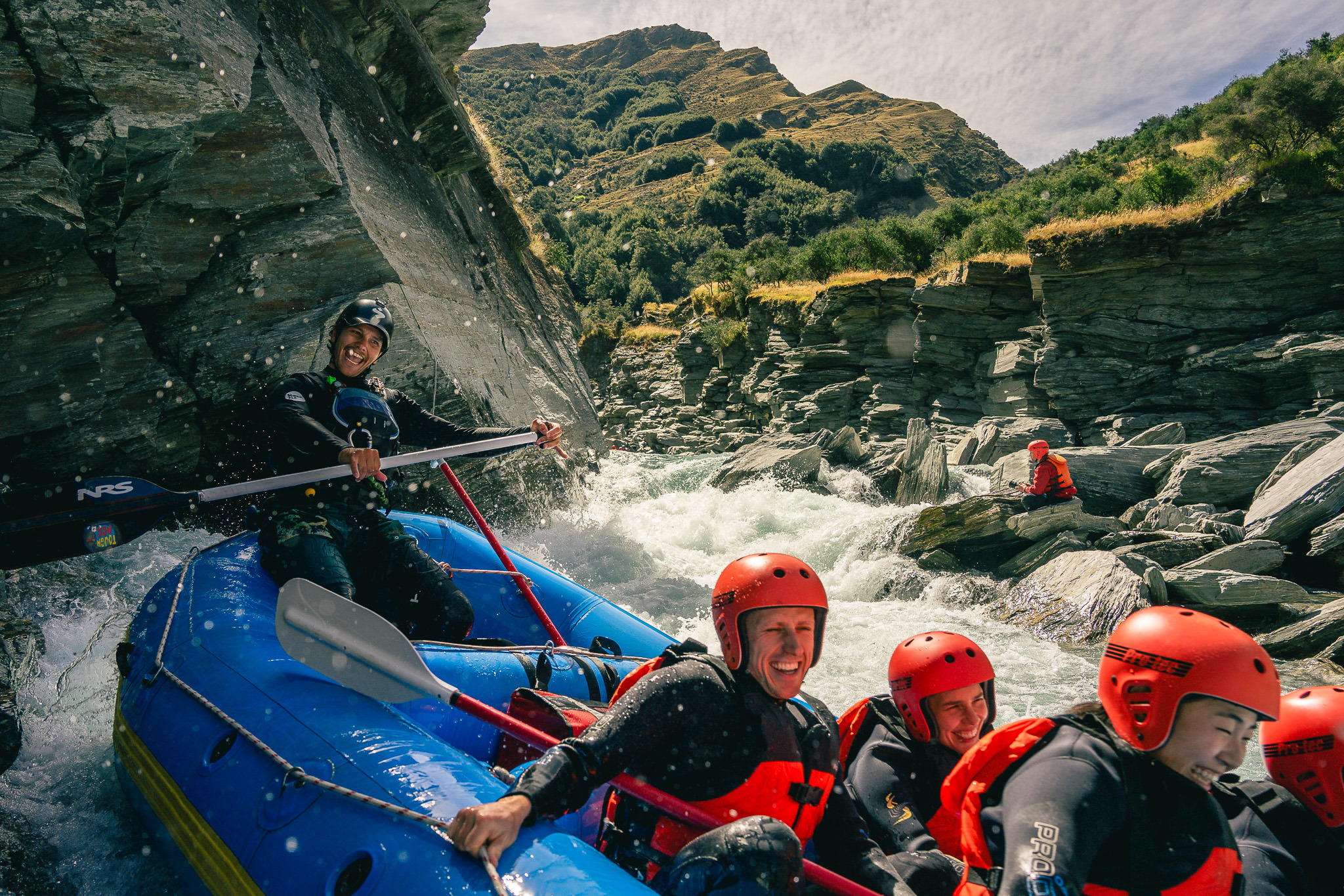 A group of smiling rafters in red helmets and wetsuits navigating white water rapids through a rocky gorge near Queenstown.