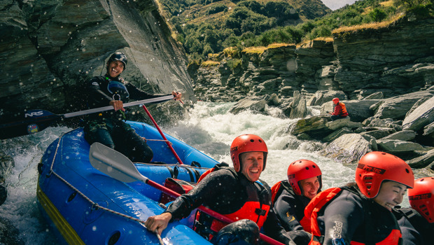 A group of smiling rafters in red helmets and wetsuits navigating white water rapids through a rocky gorge near Queenstown.