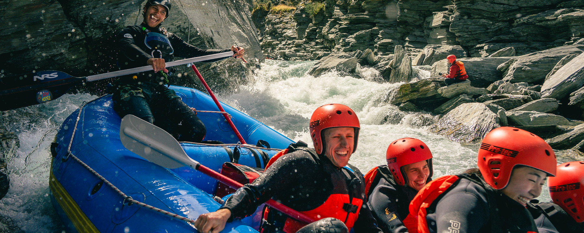A group of smiling rafters in red helmets and wetsuits navigating white water rapids through a rocky gorge near Queenstown.
