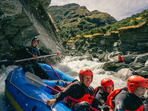 A group of smiling rafters in red helmets and wetsuits navigating white water rapids through a rocky gorge near Queenstown.