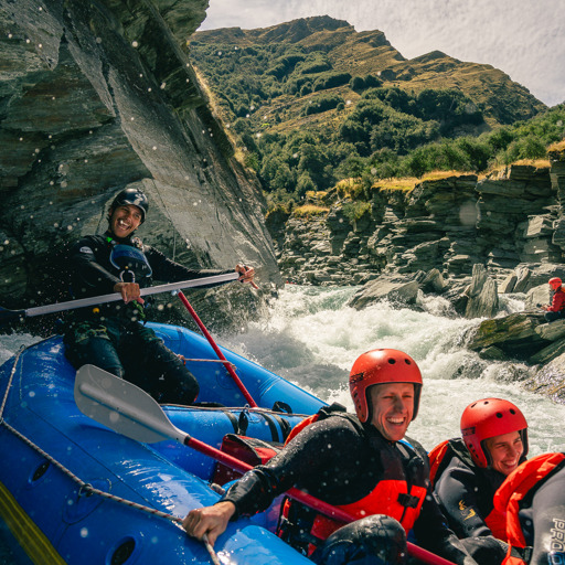 A group of smiling rafters in red helmets and wetsuits navigating white water rapids through a rocky gorge near Queenstown.