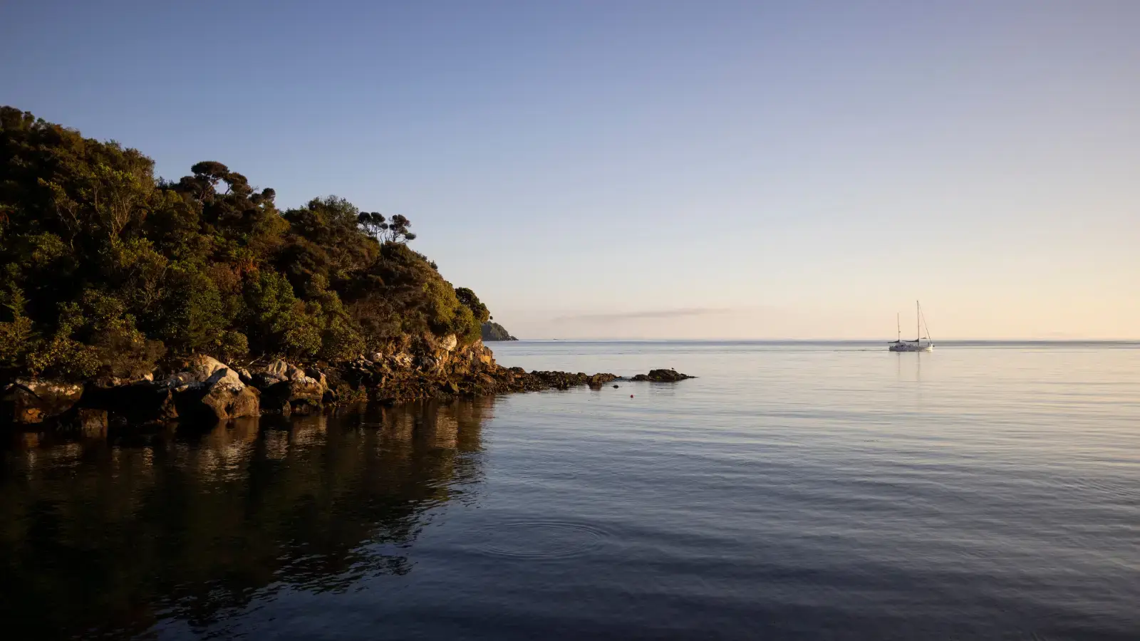 Calm coastal bay at golden hour with sailboats in the distance.