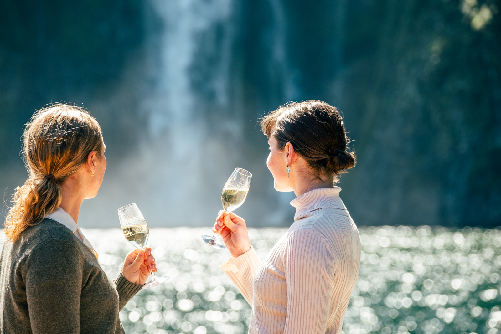Two women enjoy a glass of champagne on an outside deck of a boat while looking out at a waterfall on a sunny day. 
